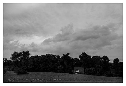 BW photograph of post-thunderstorm clouds over a meadow, treeline, and house.